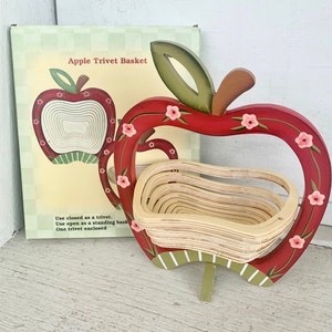 May include: A red apple-shaped trivet basket with a wooden interior. The trivet has a green stem and leaf, and is decorated with pink flowers. The box behind the trivet reads "Apple Trivet Basket".