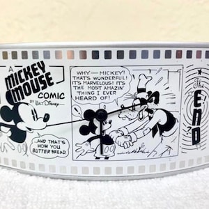 Puede incluir: Un reloj de Mickey Mouse plateado y negro con forma de carrete de película. El reloj presenta una tira cómica en blanco y negro con Mickey Mouse y Goofy. El texto de la tira cómica dice "¿Por qué - Mickey! Eso es maravilloso! ¡Es maravilloso! ¡Es lo más increíble que he oído!" y "Y así es como untas pan."