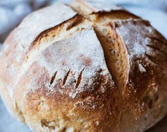 May include: A freshly baked loaf of sourdough bread with a golden brown crust and a slightly cracked surface. The bread is dusted with flour.
