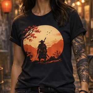 May include: Black t-shirt featuring a graphic of a samurai warrior standing in a landscape with a large orange sun. The design includes a tree with red leaves. The person is wearing black shorts and has a tattoo on their arm and leg.