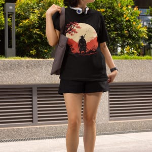 May include: Black t-shirt featuring a Japanese samurai warrior graphic. The design includes a red and beige landscape with a full moon and cherry blossom tree. The person is wearing black shorts, boots, and sunglasses.