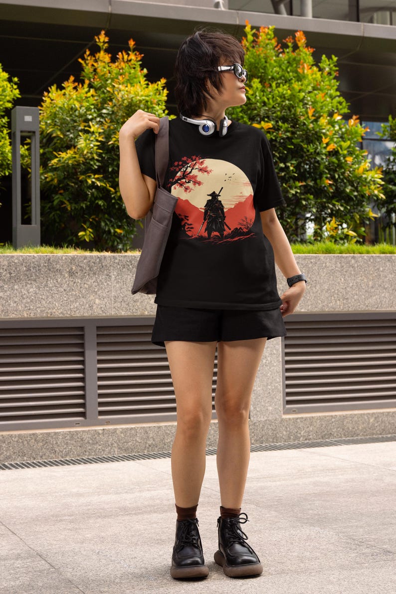 May include: Black t-shirt featuring a Japanese samurai warrior graphic. The design includes a red and beige landscape with a full moon and cherry blossom tree. The person is wearing black shorts, boots, and sunglasses.