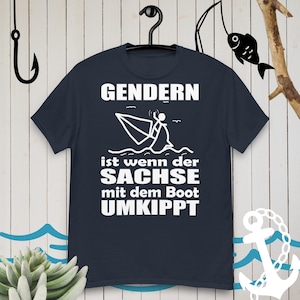 May include: A dark blue t-shirt with white text that reads "Gendern ist wenn der Sachse mit dem Boot umkippt". The text is surrounded by a simple stick figure illustration of a person in a boat that is tipping over.