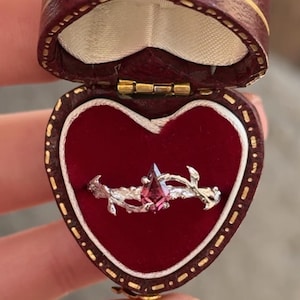 May include: A silver ring with a pink gemstone, displayed in a heart-shaped red velvet box. The ring features a floral design with a central, pointed pink stone. The box is burgundy with gold trim.