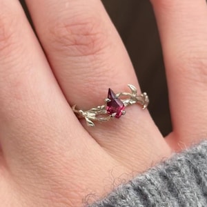 May include: A silver ring featuring a teardrop-shaped, faceted, red gemstone. The ring's band is designed with a delicate, organic, branch-like pattern. The ring is shown on a finger, with a gray sweater in the background.
