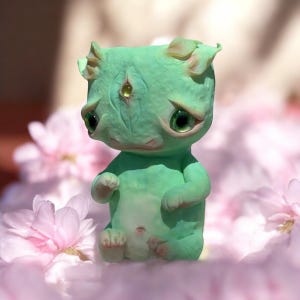 Könnte beinhalten: Eine kleine, grüne, handbemalte Figur eines Wesens mit einem dritten Auge und großen, ausdrucksstarken Augen. Die Figur sitzt auf einer rosa Oberfläche.