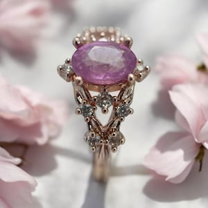 Può includere: Un delicato anello in oro rosa con una grande pietra preziosa ovale di colore viola circondata da piccole pietre preziose trasparenti.