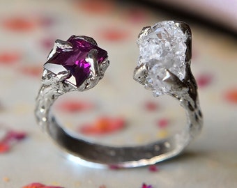May include: A silver ring with a rough, white gemstone on one side and a purple gemstone on the other. The ring is designed to look like a branch with the gemstones set in the ends.