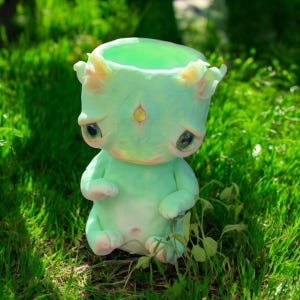 May include: A small, green, ceramic figurine with a sad expression. The figurine has large eyes, a small mouth, and a single yellow gem on its forehead. It is sitting on a bed of green grass.