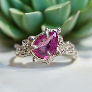 May include: A silver ring with a pink gemstone set in a claw setting. The band of the ring is textured and has a branch-like design.