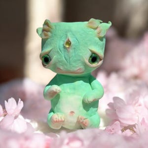 Puede incluir: Una pequeña figurita verde pintada a mano con un tercer ojo y ojos grandes y expresivos. La figurita está sentada en un lecho de pétalos de flores rosas.