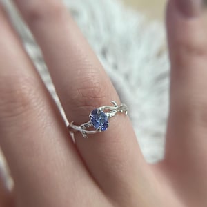 May include: A silver ring with a light blue oval gemstone. The ring's band has a delicate, twig-like design, with small branches extending around the stone. The ring is shown on a finger, highlighting its intricate details and elegant style.