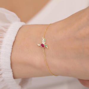 May include: A gold chain bracelet featuring a hummingbird charm. The hummingbird is decorated with red and green enamel and rhinestones.