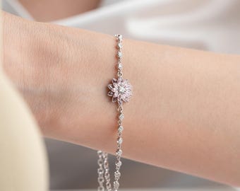 May include: A delicate silver bracelet featuring a pink flower design with a central clear stone. The bracelet has a chain with small, clear stones. The jewelry is worn on a wrist.