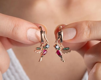Könnte beinhalten: Ein Paar goldfarbene Ohrringe mit einem Kolibri-Design, verziert mit roten, grünen und blauen Edelsteinen.