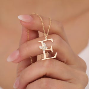 Puede incluir: Collar inicial dorado con la letra "E" y un colgante de mariposa. La mariposa está adornada con pequeños cristales brillantes. El collar está suspendido de una delicada cadena, creando un accesorio elegante y personalizado.