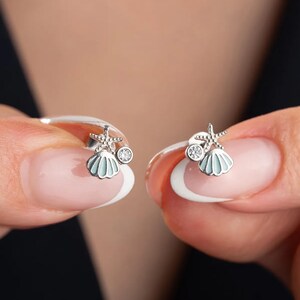 May include: A pair of silver earrings featuring a small seashell and a starfish charm. Each earring has a small clear rhinestone.