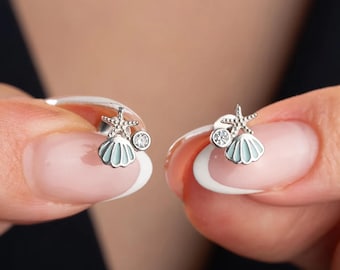 Puede incluir: Un par de pendientes de plata con una pequeña concha y un dije de estrella de mar. Cada pendiente tiene un pequeño cristal transparente.