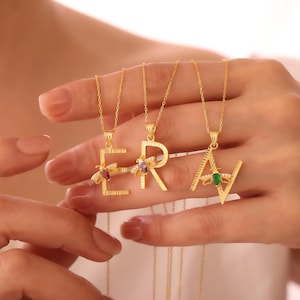 May include: Gold-colored initial necklaces spelling out the word ERA. Each letter is adorned with a small bee pendant and colorful gemstones. The necklaces are displayed against a soft background, highlighting the jewelry's intricate design and craftsmanship.