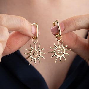 May include: A pair of gold-colored hoop earrings. Each earring features a sunburst design with a central spiral and radiating lines. The earrings are held up against a dark blue background.