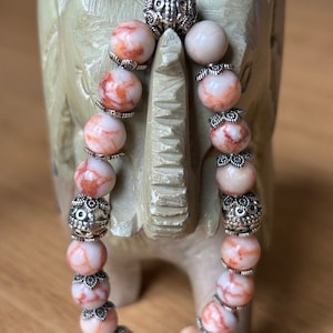 Puede incluir: Un brazalete hecho de cuentas de mármol blanco y rojo con detalles plateados. El brazalete se muestra en una pequeña figura de elefante.