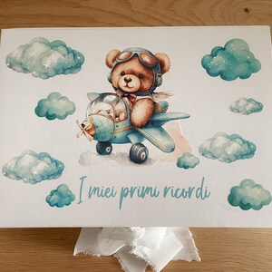 Può includere: Una scatola di legno bianca con un aereo blu e bianco e un orsacchiotto pilota. La scatola ha le parole "I miei primi ricordi" scritte su di essa.