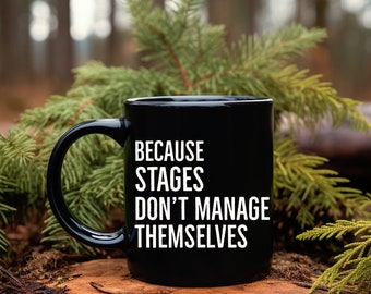 May include: Black coffee mug with white text that reads "Because stages don't manage themselves". The mug is sitting on a tree stump in a forest setting.