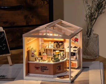 May include: A miniature dollhouse model of a bubble tea shop with a glass roof, a counter with the words "Super Bubble Tea" and a sign that says "So Cool".