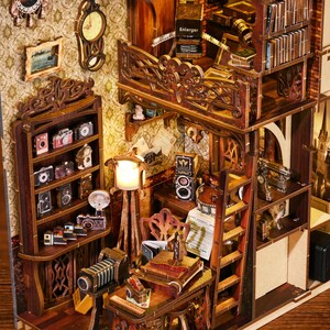 May include: A wooden miniature dollhouse with a detailed interior, featuring a bookshelf, a desk with a typewriter, a ladder, and a balcony with the word "Enlarger" printed on it.
