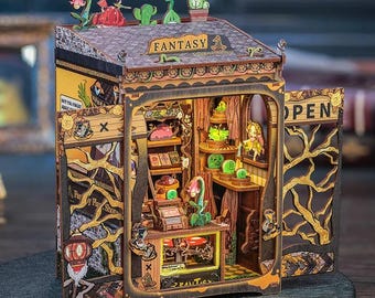 Puede incluir: Una tienda en miniatura de madera con un letrero que dice "Fantasy" y una puerta abierta que dice "Open". La tienda está decorada con plantas, flores y una pequeña figura de una persona. La tienda está hecha de madera y tiene una combinación de colores marrón y dorado.