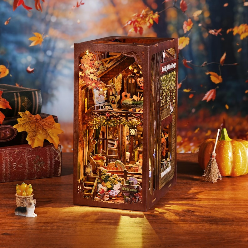 May include: A wooden, illuminated book nook diorama with a cozy autumn theme. The miniature scene includes a house, trees, and fall foliage. The text "Twilight Cottage" is visible. A pumpkin and broom are on the table.