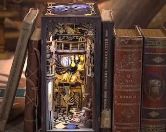May include: A miniature wooden model of a book with a detailed interior scene. The book is open to reveal a small room with a desk, chair, and bookshelves. The room is lit by a lamp and has a window overlooking a forest. The book is surrounded by other books.