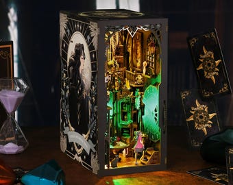 Könnte beinhalten: Eine Holzkiste mit einer Miniatur-Szene im Inneren. Die Kiste ist mit einem schwarzen und goldenen Design verziert und zeigt eine Frau mit langen schwarzen Haaren. Die Szene in der Kiste ist mit grünen Lichtern beleuchtet und zeigt eine kleine Figur, die auf einer Treppe steht.
