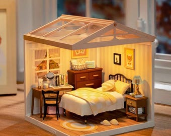 May include: A miniature dollhouse bedroom with a white bed, a wooden dresser, a desk with a chair, and a bedside table with a lamp. The room has a glass roof and is lit up with warm white lights.