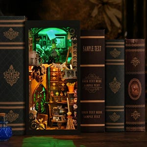 May include: A miniature dollhouse with a green lit interior, featuring a witch's workshop with a cauldron, broom, and shelves filled with potions. The dollhouse is displayed on a wooden surface with books behind it.
