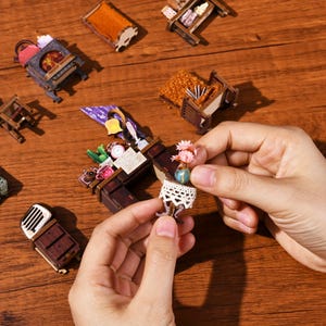 May include: Miniature wooden furniture pieces, including a fireplace, desk, and bed, are scattered on a wooden surface. A small table with a lace doily and a vase of pink flowers is held by a person's hands. The scene evokes a dollhouse or craft project.