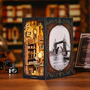 May include: A miniature wooden model of a shop with a window display featuring a clock and other items. The shop is open and the interior is visible. The model is set against a backdrop of a black and white photo of the Tower Bridge in London. The text "The beautiful moments freeze in the moment of gaze" is printed on the model.