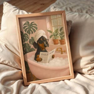 May include: A framed print depicts a dachshund in a bathtub, wearing a towel and glasses, reading a newspaper labeled "DAILY NEWS." The artwork has a soft, pastel color palette, with plants and a window in the background.