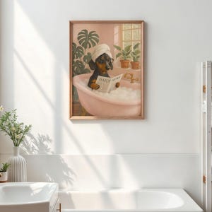 May include: A framed print depicts a dachshund in a pink bathtub, wearing a towel and glasses, reading a newspaper. The print has a wooden frame and is set against a pink wall with plants.