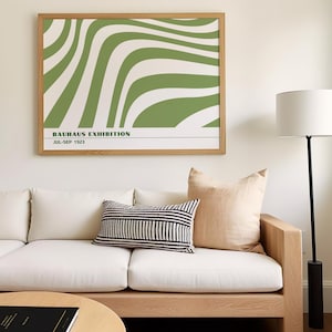 May include: A framed Bauhaus Exhibition poster with a green and white wavy design hangs above a white sofa with decorative pillows. The poster's text reads "BAUHAUS EXHIBITION JUL-SEP 1923". A floor lamp with a white shade stands to the right.