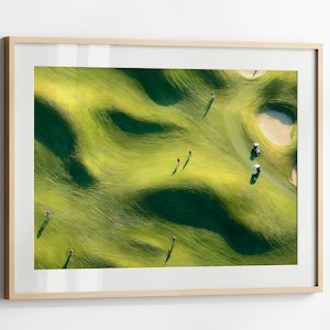 Puede incluir: Vista aérea enmarcada de un campo de golf con calles verdes onduladas y una trampa de arena. Se ven varios golfistas y carritos de golf. La imagen está enmarcada en un marco de madera clara.