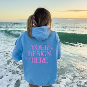 May include: A light blue hooded sweatshirt with the text "YOUR DESIGN HERE" in pink lettering on the back. The sweatshirt is worn by a person standing on a beach with white sand and blue water.
