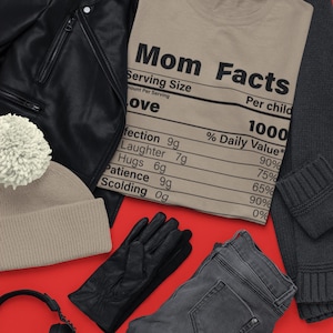 May include: A flat lay featuring a brown t-shirt with the text "Mom Facts" and a nutrition label listing "Affection, Laughter, Hugs, Patience, Scolding" with percentages. The t-shirt is surrounded by a black leather jacket, a grey sweater, a pair of grey jeans, black leather gloves, a beige beanie with a white pom-pom, a pair of black headphones, and a black travel mug.