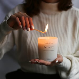 Può includere: Una persona accende una candela bianca con la parola "Serenity" stampata sul davanti. La candela è in un barattolo di vetro trasparente.
