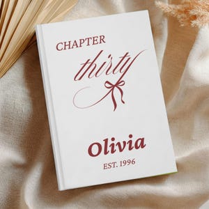 May include: A white hardcover book with dark red text that reads CHAPTER thirty with a small bow graphic, followed by the name Olivia and the text EST. 1996. The book cover features a clean design with centered typography and a minimalist aesthetic.