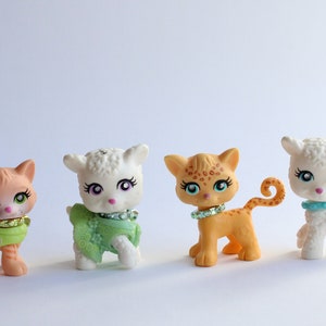 Könnte beinhalten: Vier kleine Tierfiguren aus Plastik, zwei weiße Lämmer, eine braune und orange Katze mit einem grün-rosa Halsband und eine braune und orange Katze mit einem weiß-blauen Halsband.