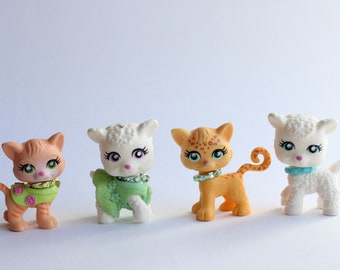 May include: Four small plastic animal figurines, two white lambs, a brown and orange cat with a green and pink collar, and a brown and orange cat with a white and blue collar.