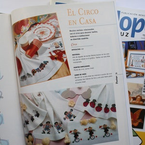 May include: A colorful embroidery pattern for a circus-themed kitchen towel. The pattern features a red, white, and blue striped tent, a clown riding a unicycle, and a red apple. The text "El Circo en Casa" is printed on the page.