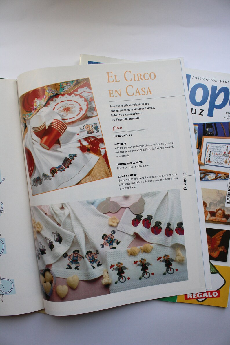 May include: A colorful embroidery pattern for a circus-themed kitchen towel. The pattern features a red, white, and blue striped tent, a clown riding a unicycle, and a red apple. The text "El Circo en Casa" is printed on the page.