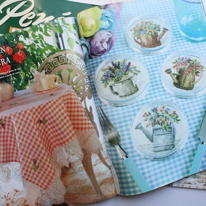 May include: A magazine spread featuring a cross-stitch pattern for a set of four coasters. Each coaster features a watering can filled with flowers. The magazine is open to a page with a blue and white checkered background and the text "ION MENSUAL N 64".